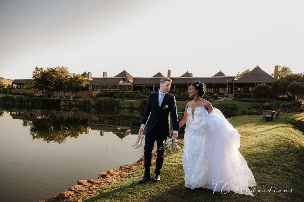 A man and woman in wedding attire holding hands and walking by water at kloofzicht lodge & Spa